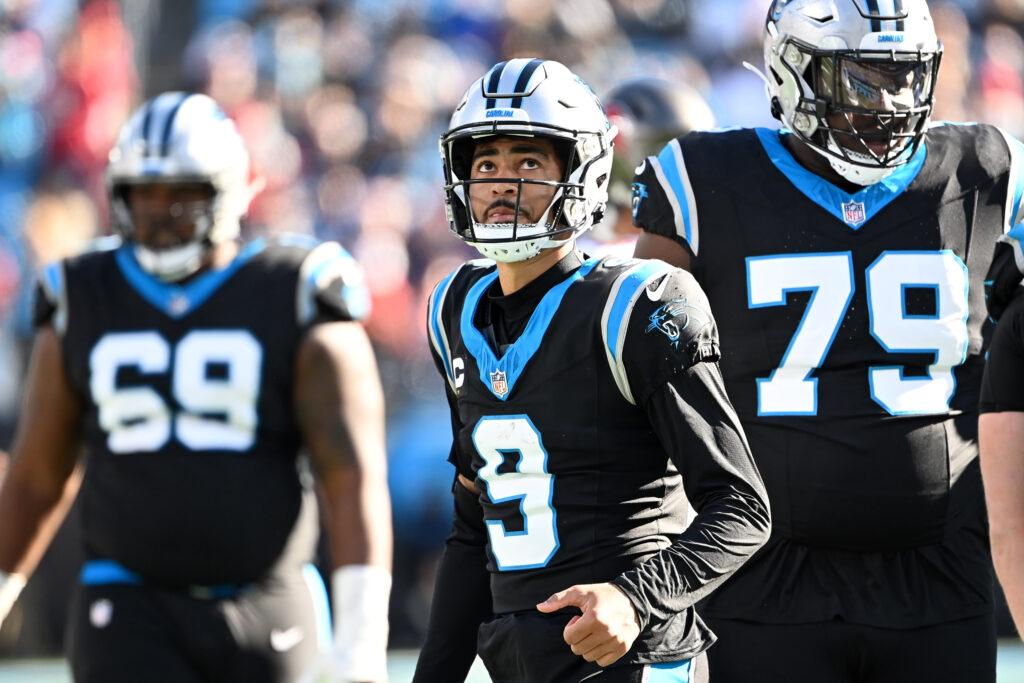 Zoomed in image of Carolina Panthers QB Bryce Young (black jersey, silver helmet; blue accents on both) looking up at the scoreboard