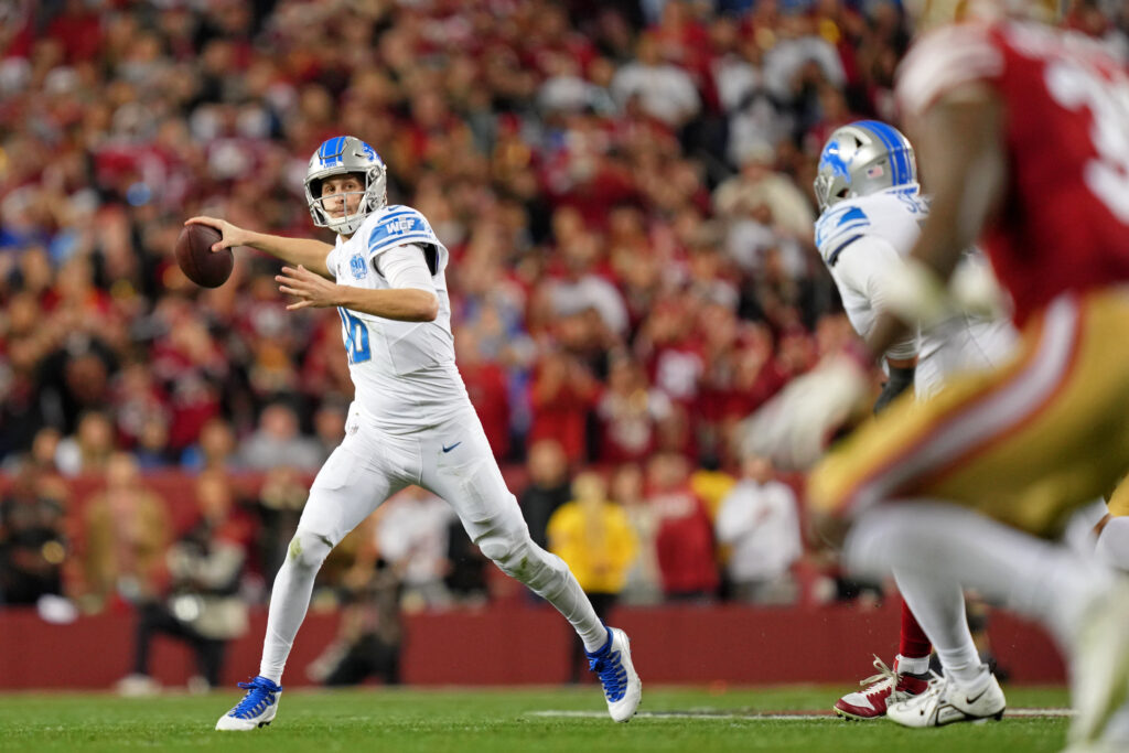 Detroit Lions QB Jared Goff (white jersey, pants and cleats; silver helmet; all with blue accents) gets ready to throw the ball in space