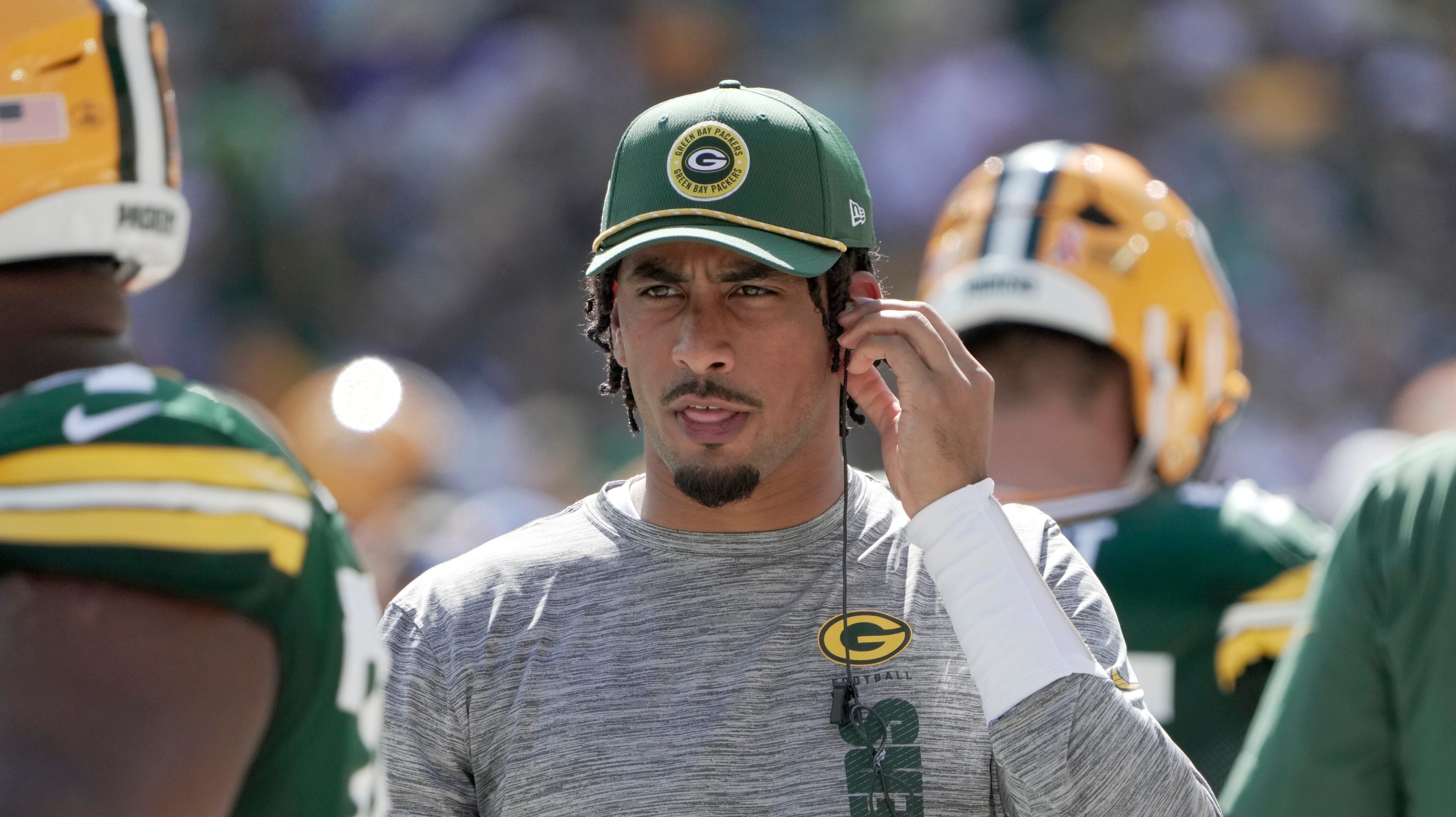 Jordan Love stands on the sideline in street clothes reaching for his ear piece.